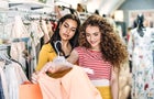 Two female teenager friends standing inside in the shop, holding and looking at dress. 